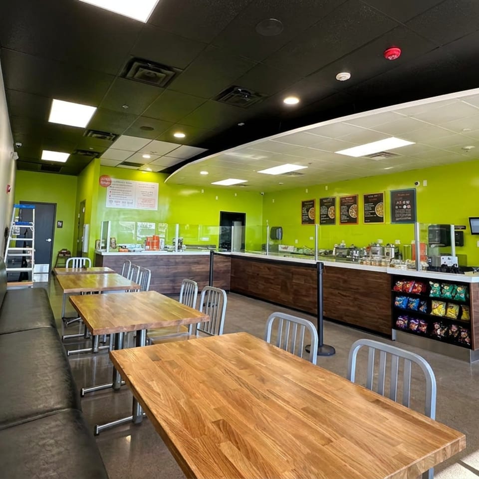 Bright casual restaurant interior with wooden tables, metal chairs, booth seating, lime-green walls, service counter, menu boards, and Pepsi refrigerator.