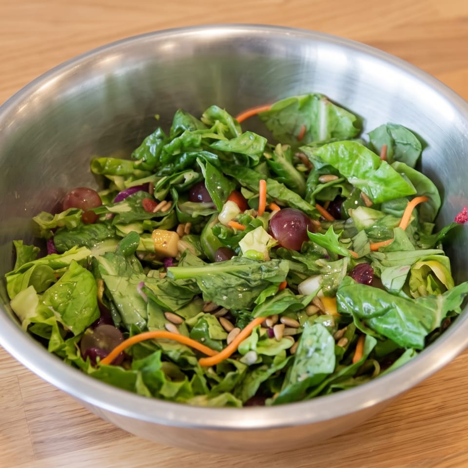 Fresh green salad with grapes, shredded carrots, cheese cubes, and seeds in a metal bowl on a wooden table.